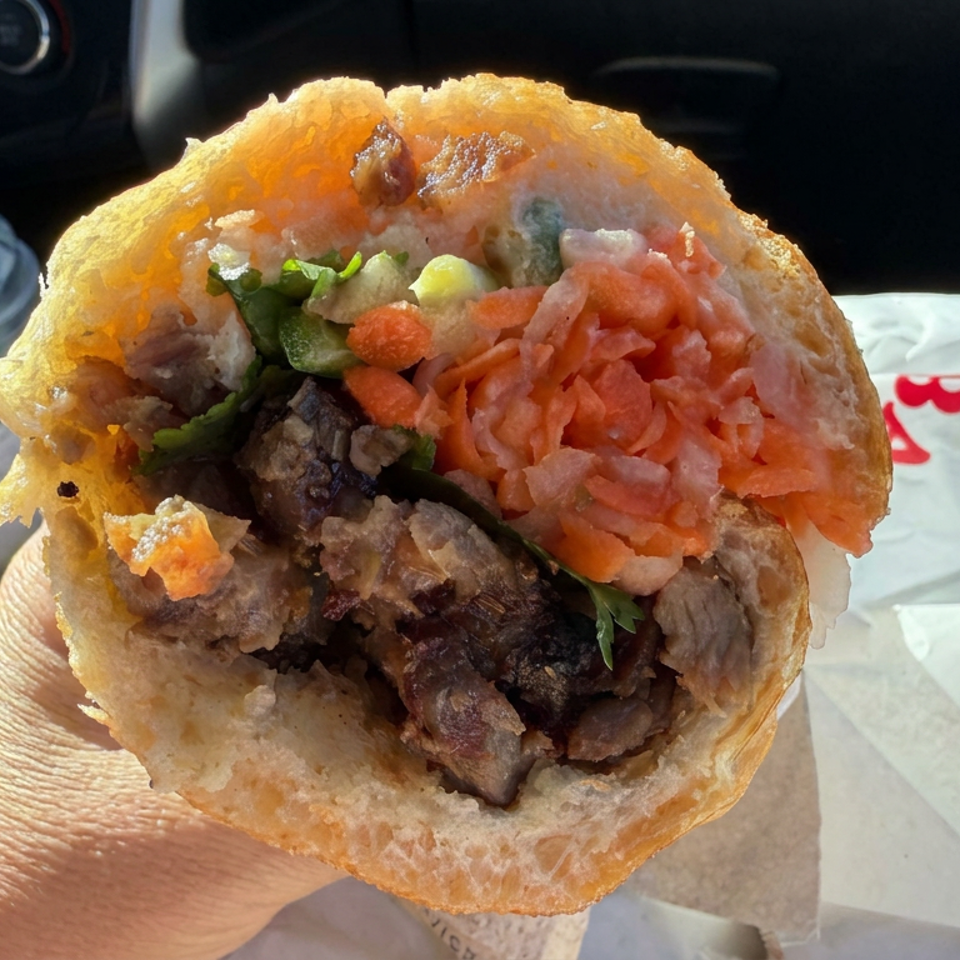 Close-up of a stuffed pita sandwich with grilled meat, shredded vegetables, and herbs, held inside a car.