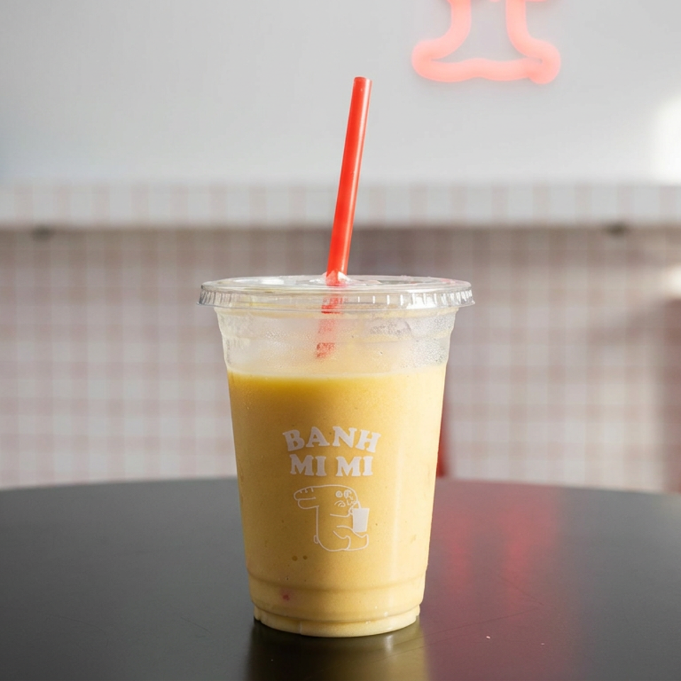 Iced yellow drink in a branded Banh Mi Mi cup with a red straw on a café table.