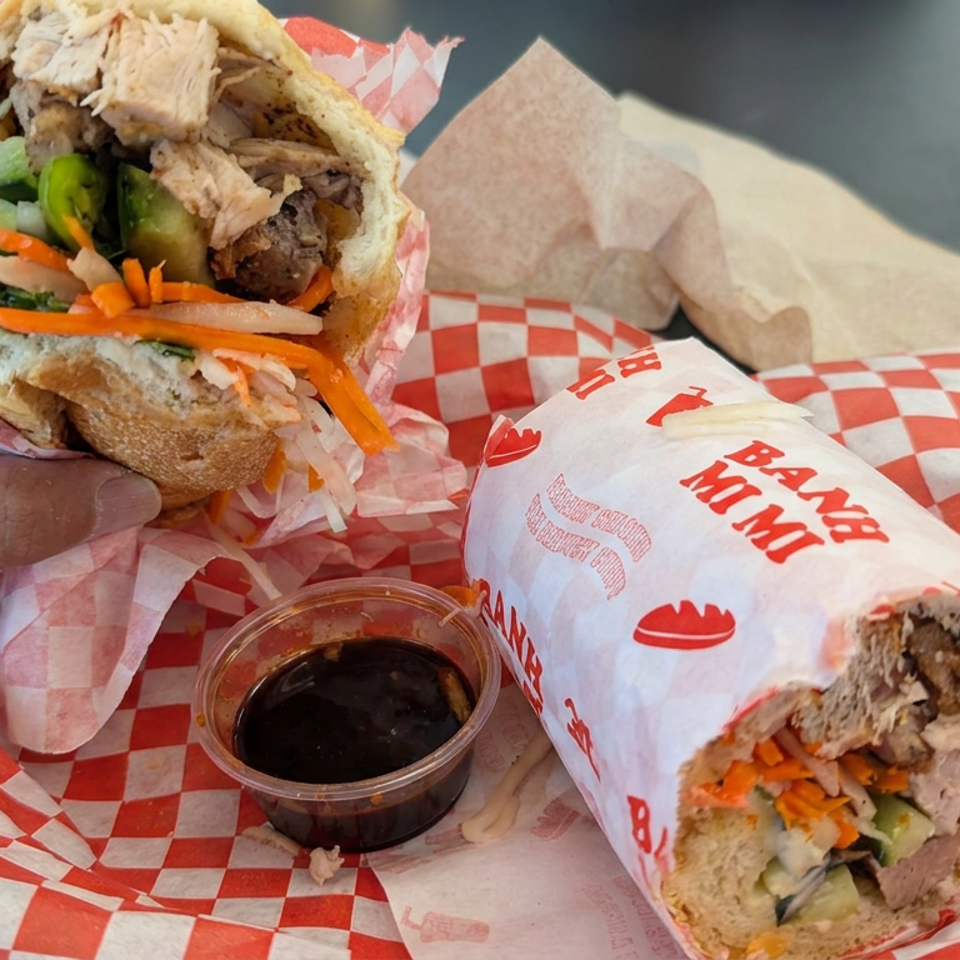 A hand holds a stuffed pita wrap with roasted meat and vegetables beside a wrapped sandwich and dipping sauce.
